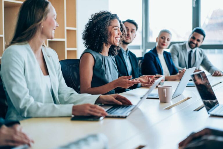 A large group of office workers seated in a boardroom all turn to face a female colleague who is smiling and gesturing animatedly as she talks.