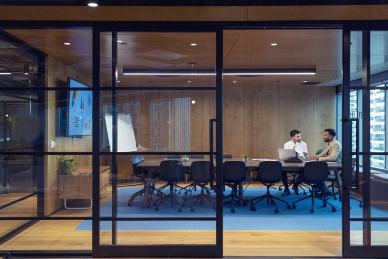 Two male colleagues sit in a large glass fronted board room.