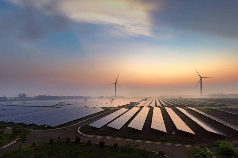 The sun rises over a field of solar panels with two wind turbines standing in the middle distance.