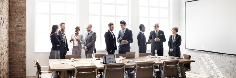 A group of office workers stand deep in conversation behind a table in a large board room table.