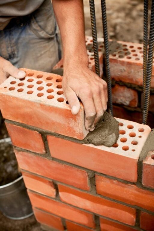 A builder places a red brick onto the wall he is building.