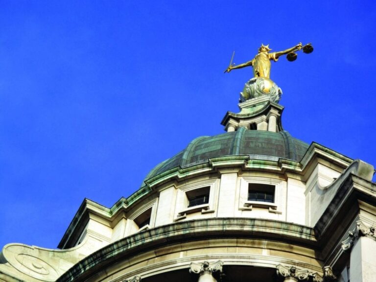 A view from below of a golden statue brandishing a sword and scales atop a building, framed by the vibrant blue of the sky in the backdrop.