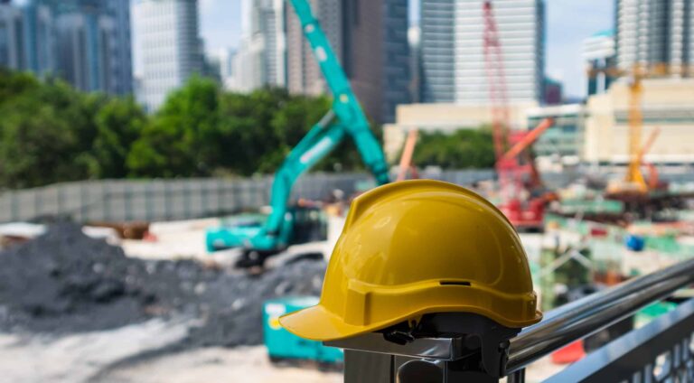 A yellow hard hat sits on of a fencepost on a construction site while cranes work in the back ground.