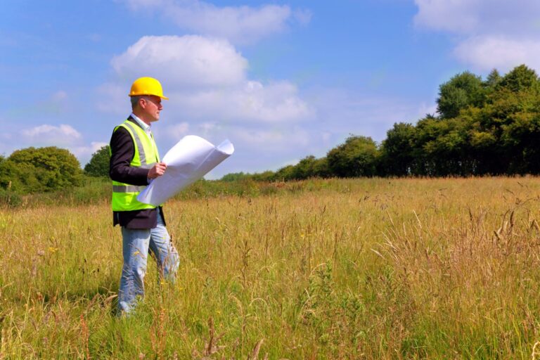 A construction worker, clad in a hard hat and high-visibility vest, stands in a green field, holding architectural plans.