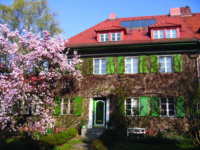 A large red roofed, and green shuttered property stands half shaded by a magnolia tree in a country garden.