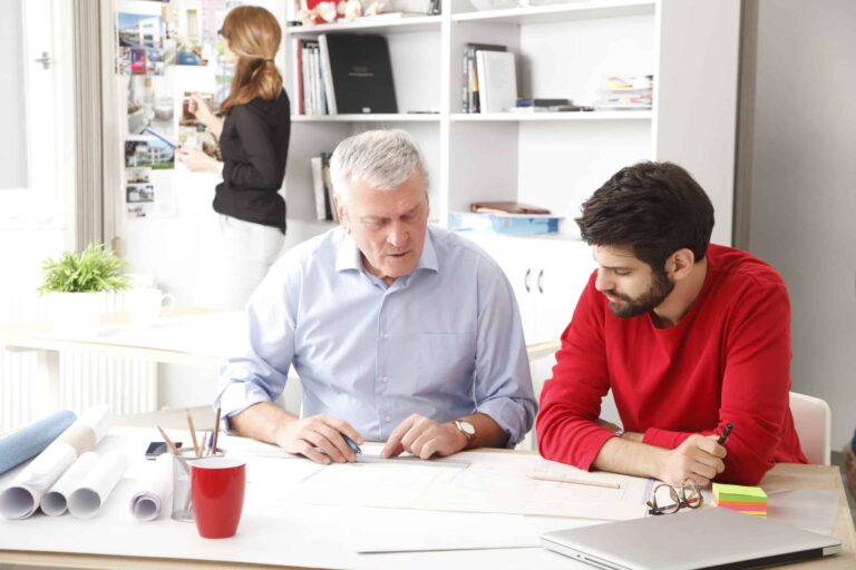 A father and son sit at a desk strewn with building plans and look deep in conversation while a woman peruses photographs pinned to a wall.