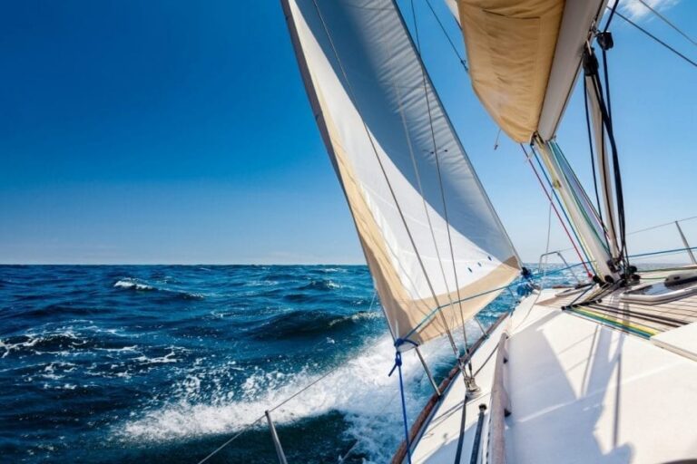 A sail boat glides through the waves and tilts to one side as the wind fills its sails, the clear blue sky as its backdrop.