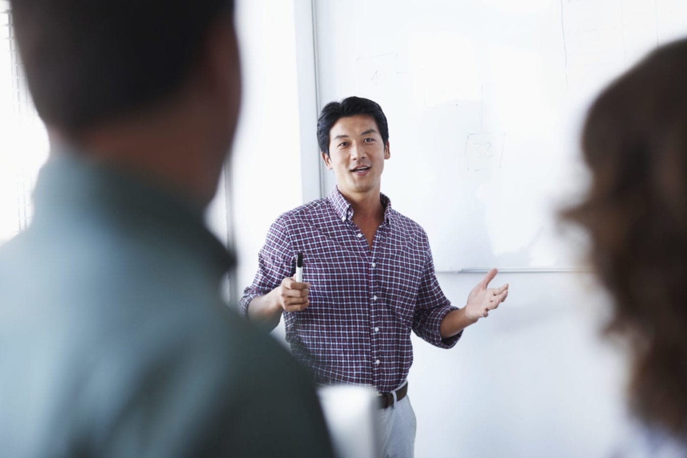 A man stands in front of a whiteboard, holding a black marker pen, presenting to his colleagues.