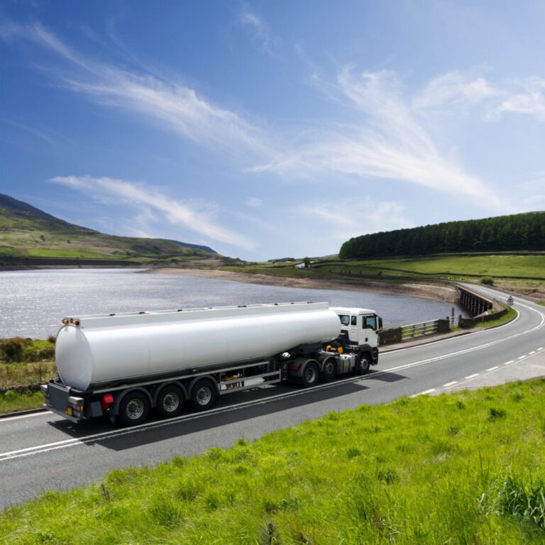 An oil truck makes its way down a deserted road, approaching a bridge that spans a lake, bathed in the sunlight.