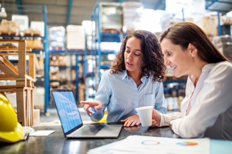 Two women sat at a desk inside a warehouse looking at figures displayed on a laptop computer.