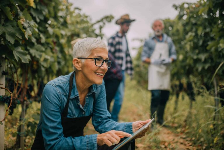 The female business owner sits looking at an I-Pad screen in her vineyard, while two male workers converse in the background.