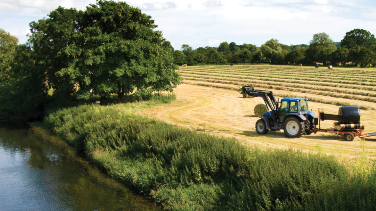 A blue tractor in a field on the right, next a river on the left. It is a bright, cloudy day.