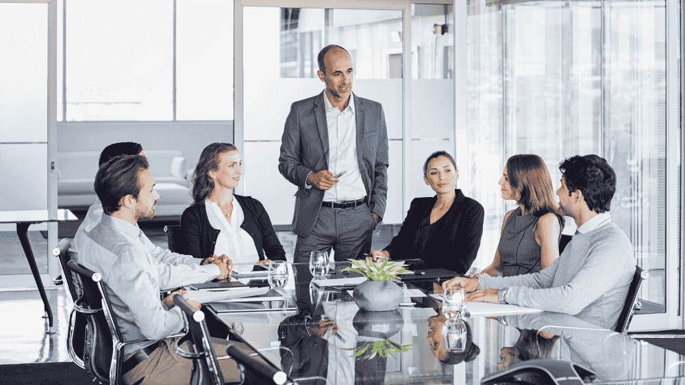 business people sit around a table and one, a man stands talking, they are in a modern office and all wearing smart suits