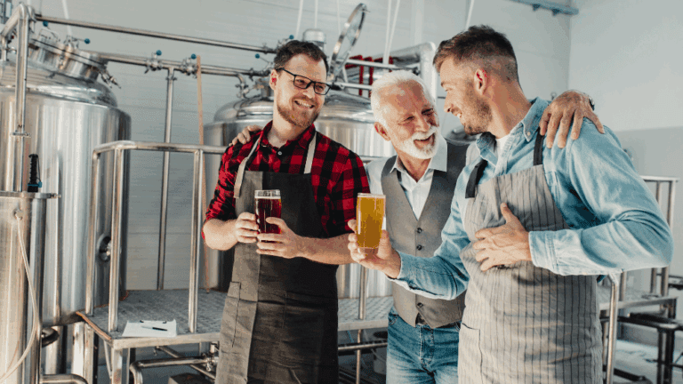A family business owner with his two adult sons. The man has his arms around his two adult sons who are holding a pint of cider each. They are all smiling and chatting. The setting is in a brewery.