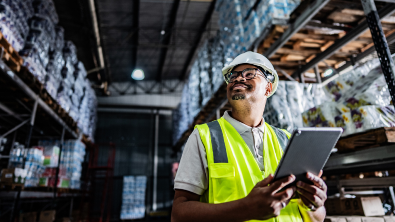 Warehouse operative using a tablet to manage inventory in a logistics warehouse, surrounded by stocked pallets and shelving, representing supply chain and international trade operations.